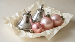 Nature morte composée de cloches et de boules de Noël en verre soufflé des années 1930, posées sur un papier de soie ivoire dans une douce lumière hivernale, aux teintes d’argent patiné et de rose nacré.